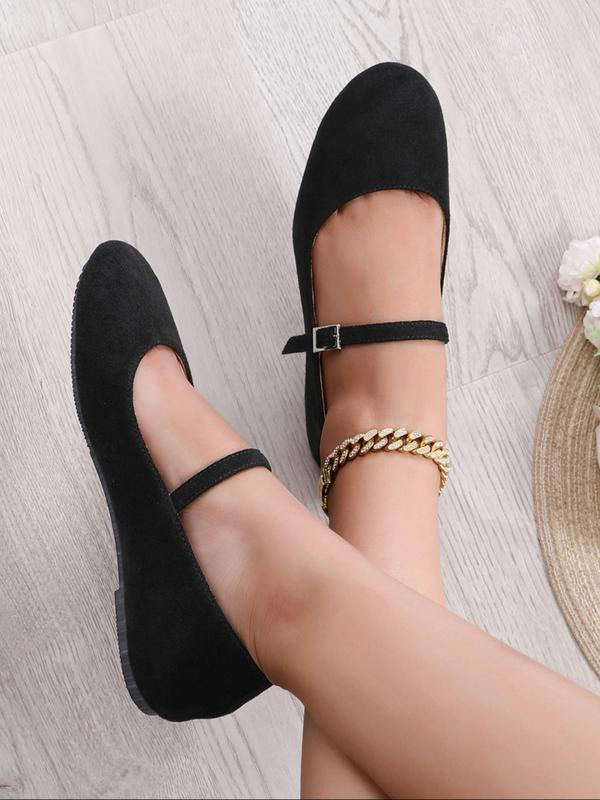 Black flat shoes worn with a gold chain bracelet on a light wooden floor.
